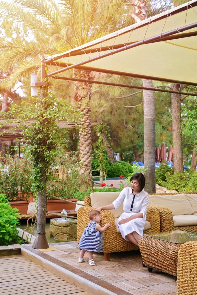 fashionable mother spending time with her little daughter at the stylish wooden terrace with wicker garden furniture at sunny summer day